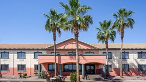 A hotel with a two-story facade, a covered patio area with tables and chairs, and palm trees in front. - Baymont by Wyndham Phoenix I-10 near 51st Ave (Phoenix)