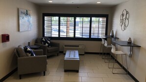 A modern living room with a sofa, ottoman, and a coffee table. There is a large window with blinds, a painting on the wall, and a small red object on the left. - Baymont by Wyndham Phoenix I-10 near 51st Ave (Phoenix)