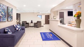 A reception area with a blue carpet, a sofa, two chairs, a small table with a vase, and a monitor on a stand. - Baymont by Wyndham Phoenix I-10 near 51st Ave (Phoenix)