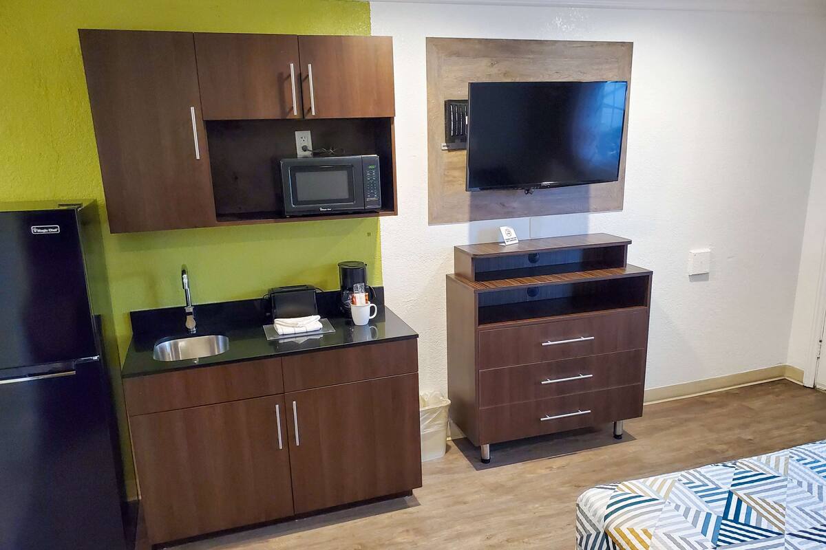 Une cuisine compacte équipée d’un four à micro-ondes, d’une machine à café et d’un évier. On y trouve un lit avec une couvre-lit à motifs et un téléviseur fixé au mur.