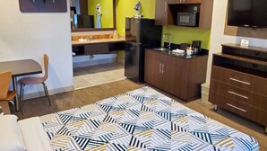 Habitación de hotel compacta con cama, escritorio, kitchenette y televisor.