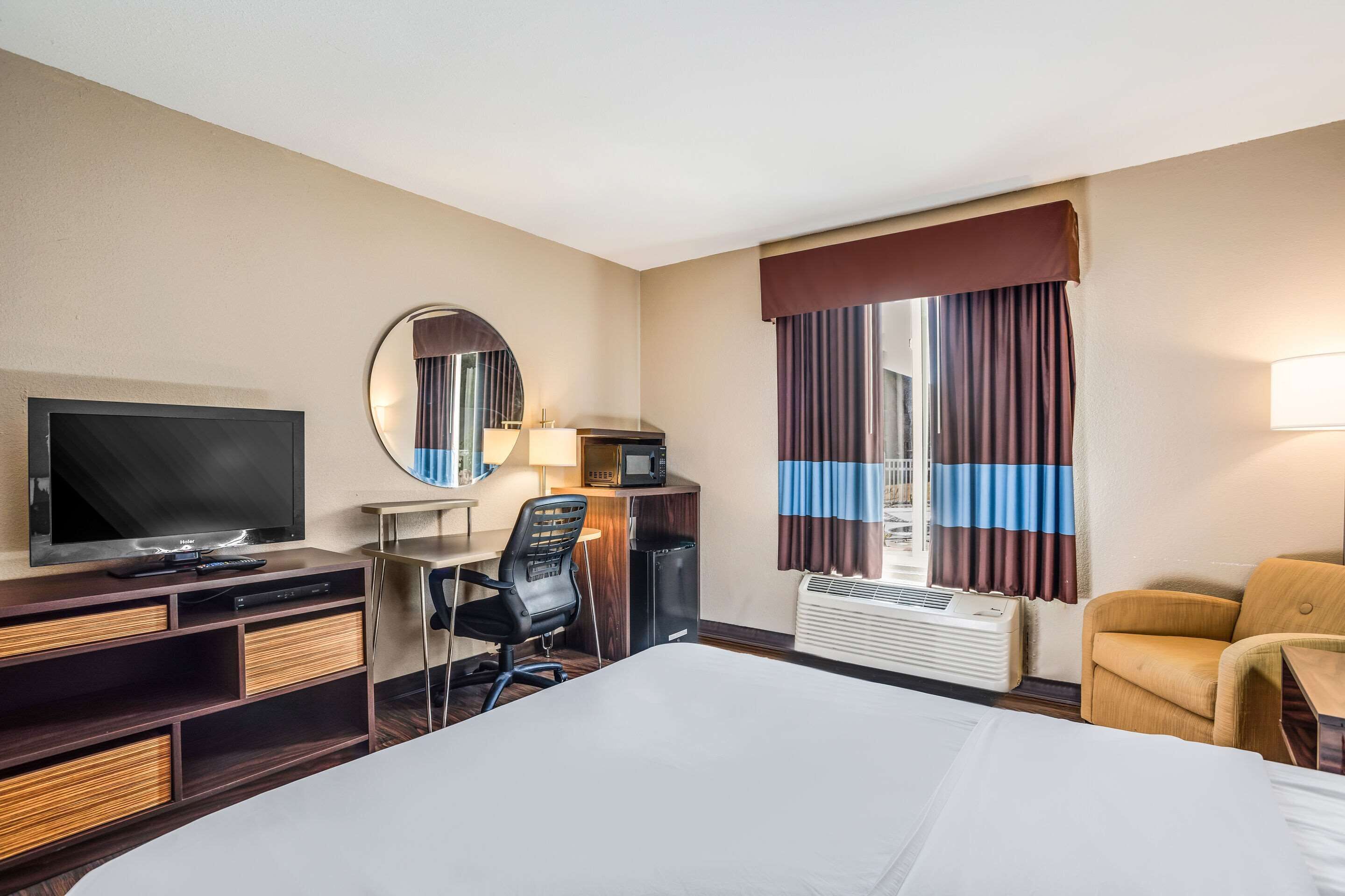 Una habitación de hotel con una cama grande, un escritorio con silla, televisión, espejo, calentador y una ventana con cortinas.