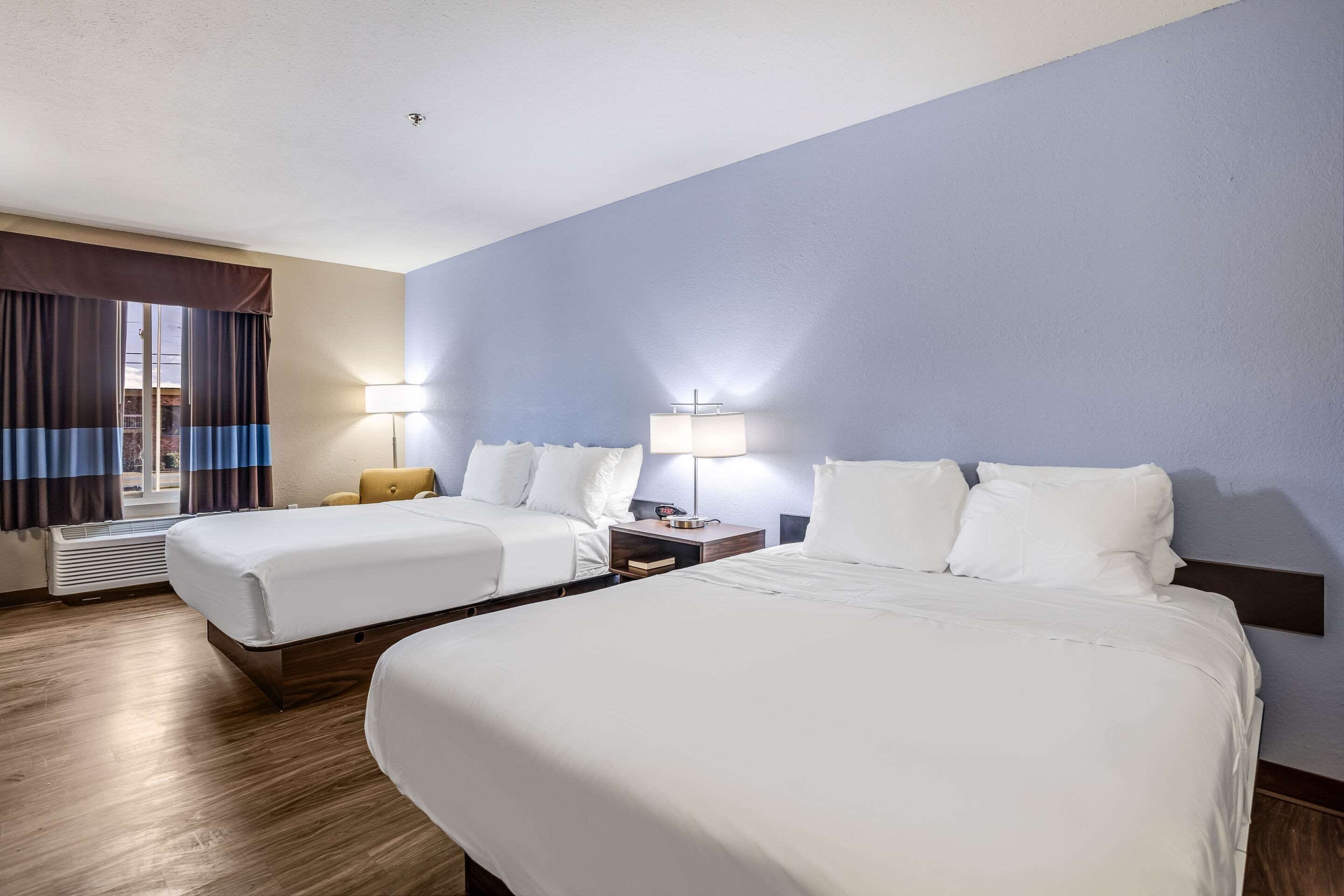 Habitació d'hotel amb dos llits, una tauleta de nit amb una làmpada, una finestra amb cortines i un aparell d'aire condicionat fixat a la paret.