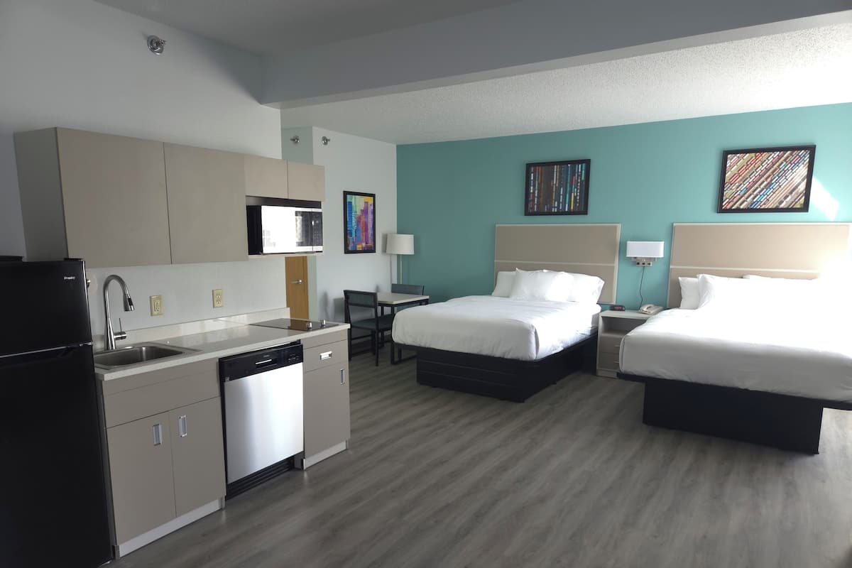 A hotel room with a kitchenette, two beds, a dining table, and artwork on the walls.