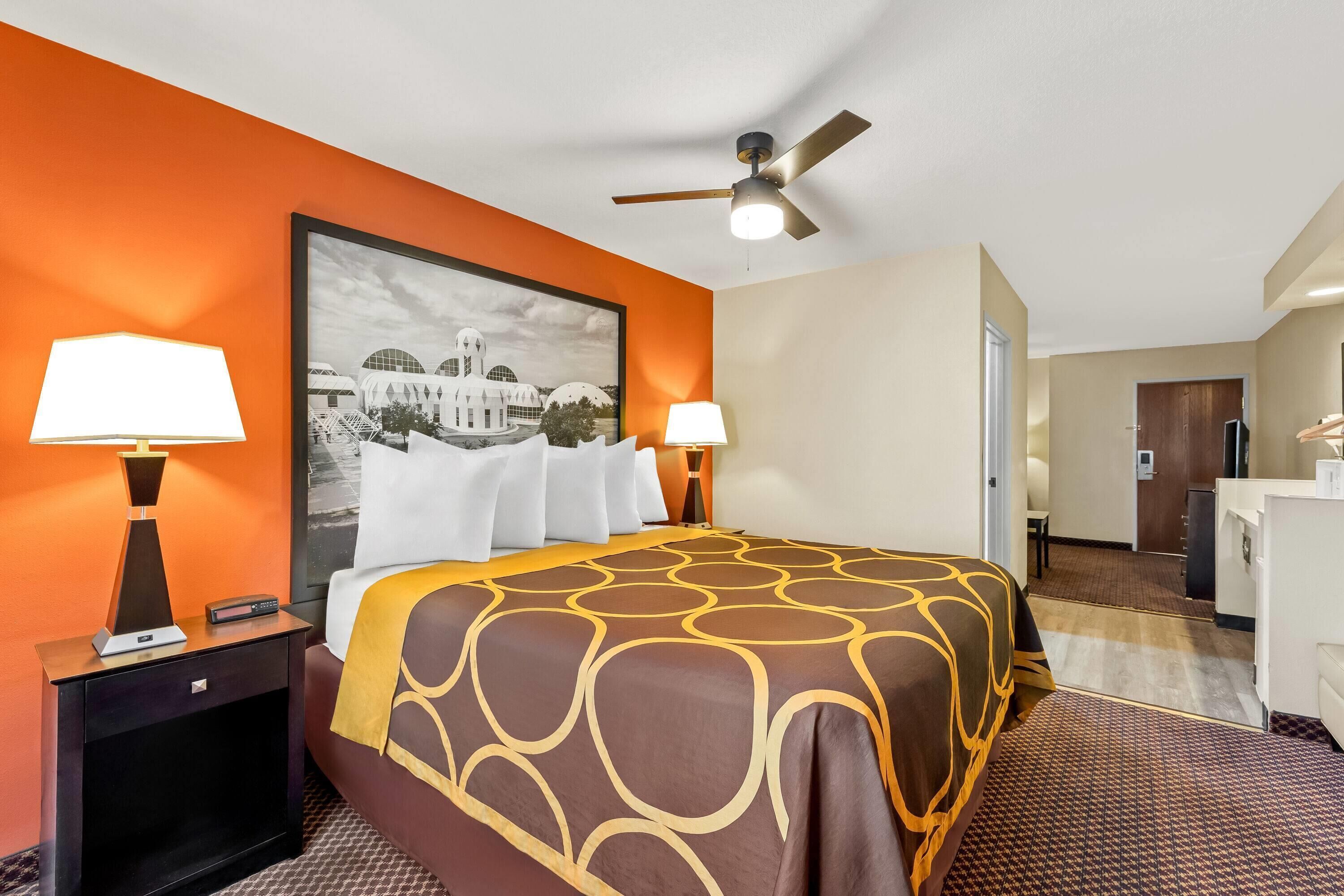 A hotel room with an orange accent wall, a bed with a patterned bedspread, two bedside lamps, a ceiling fan, and a framed picture on the wall.