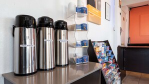Un bancone con tre macchine da caffè in acciaio inox, etichettate con “ACQUA CALDA” e “Caffè DECEDORATO”, un espositore per caffè e un porta riviste con diverse pubblicazioni.