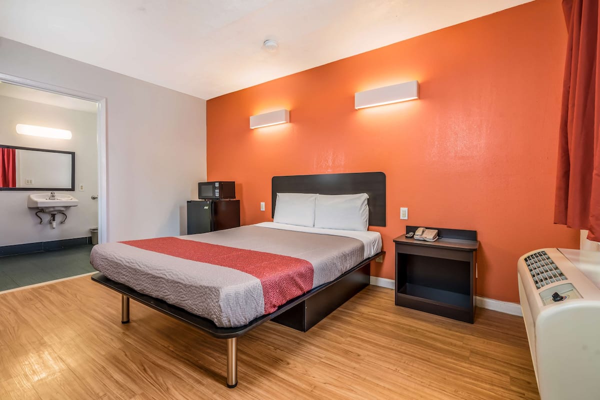 A hotel room with a bed, bedside tables, a microwave, a heater, and a bathroom with a sink and mirror.