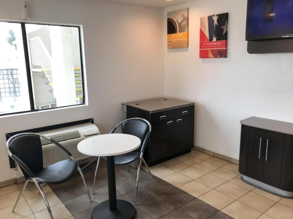 A small, modern room with a round table and two chairs, a cabinet with drawers, a radiator, and a window with a view of a building outside.