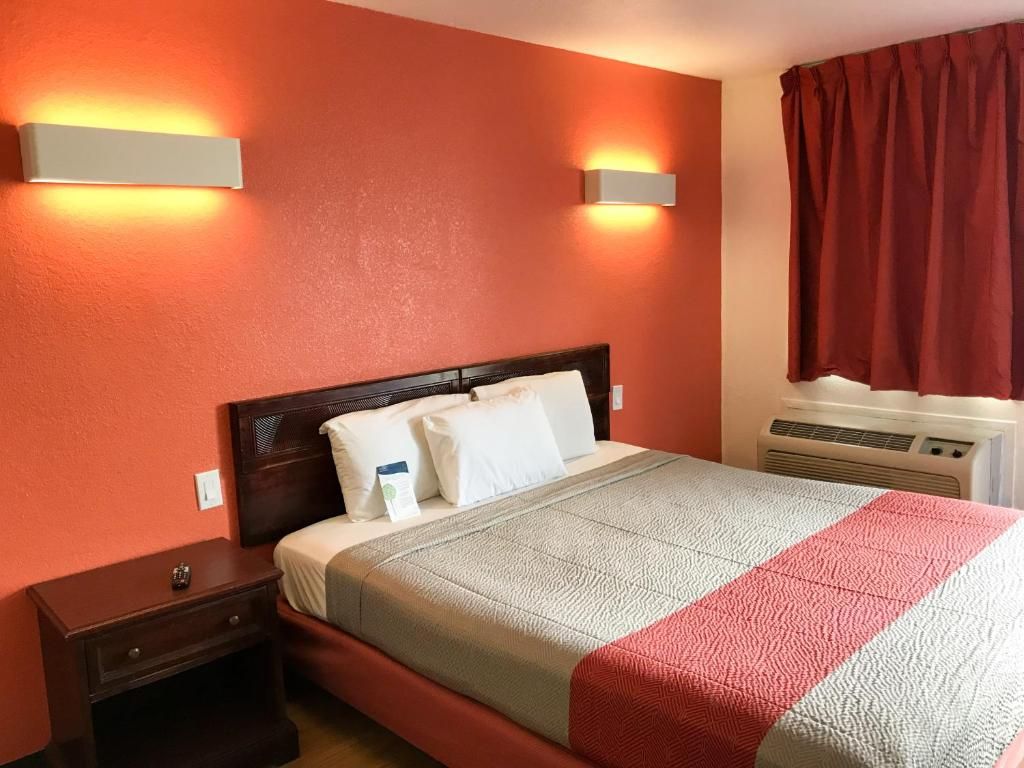 A hotel room with a bed, nightstand, wall-mounted light fixtures, and an air conditioning unit.