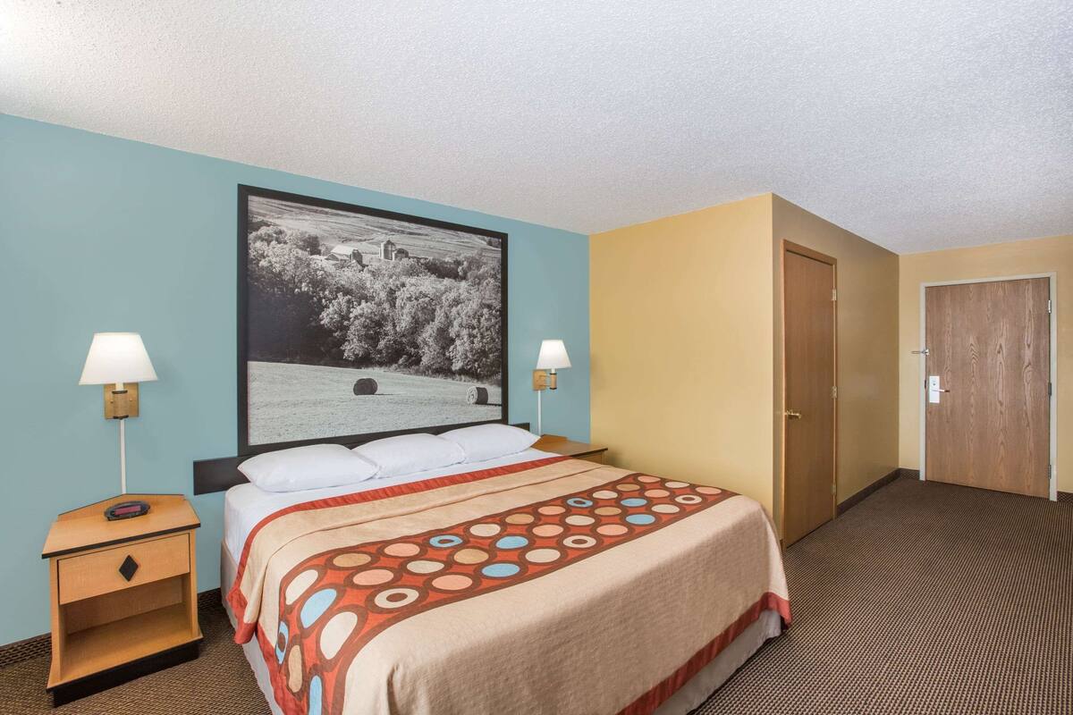 Une chambre d’hôtel avec un lit, deux lampes de chevet, une photographie en noir et blanc encadrée accrochée au mur, et une porte.