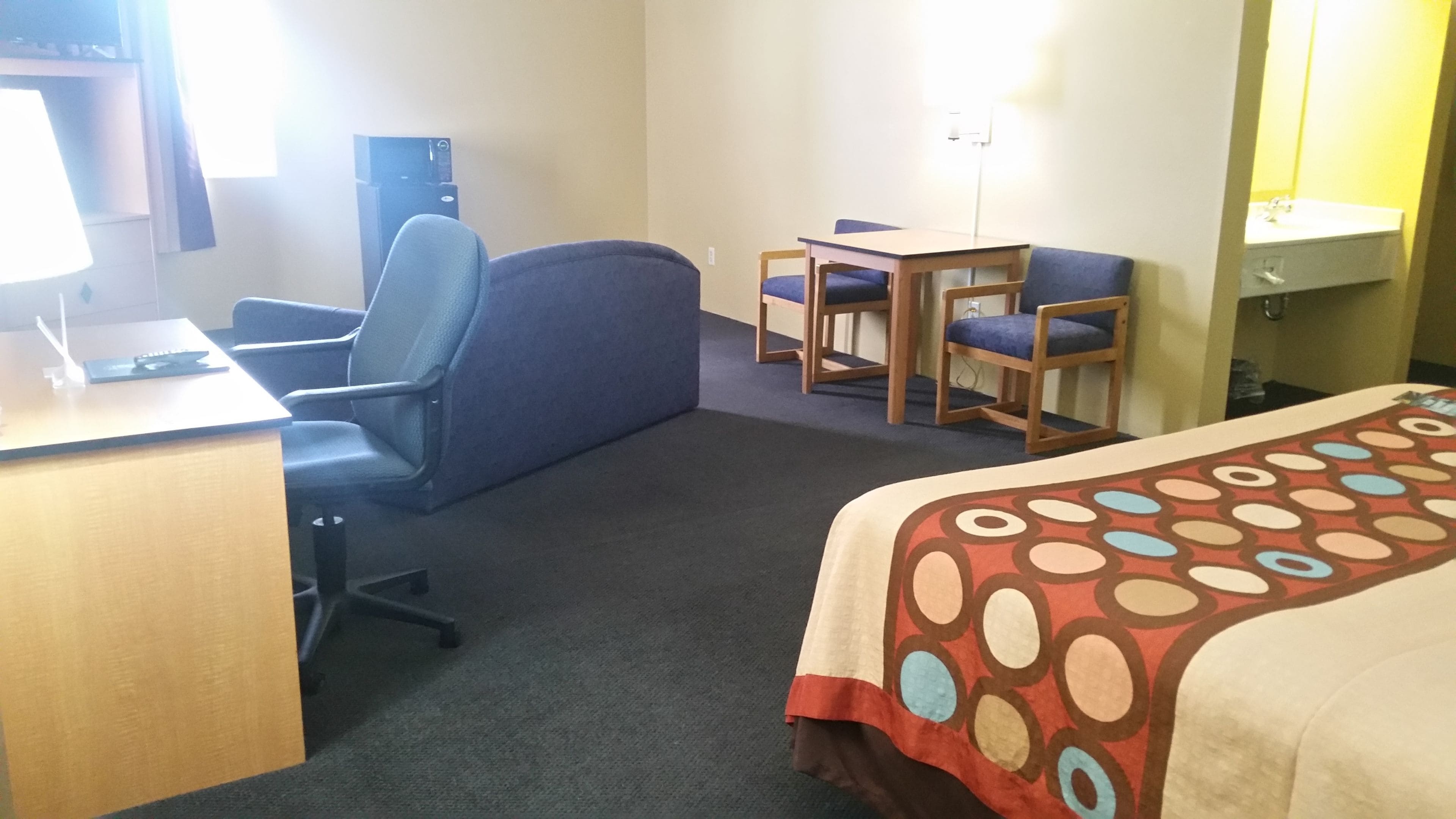 A hotel room with a desk, office chair, two chairs, a bed with a patterned bedspread, a small table, and a mirror.