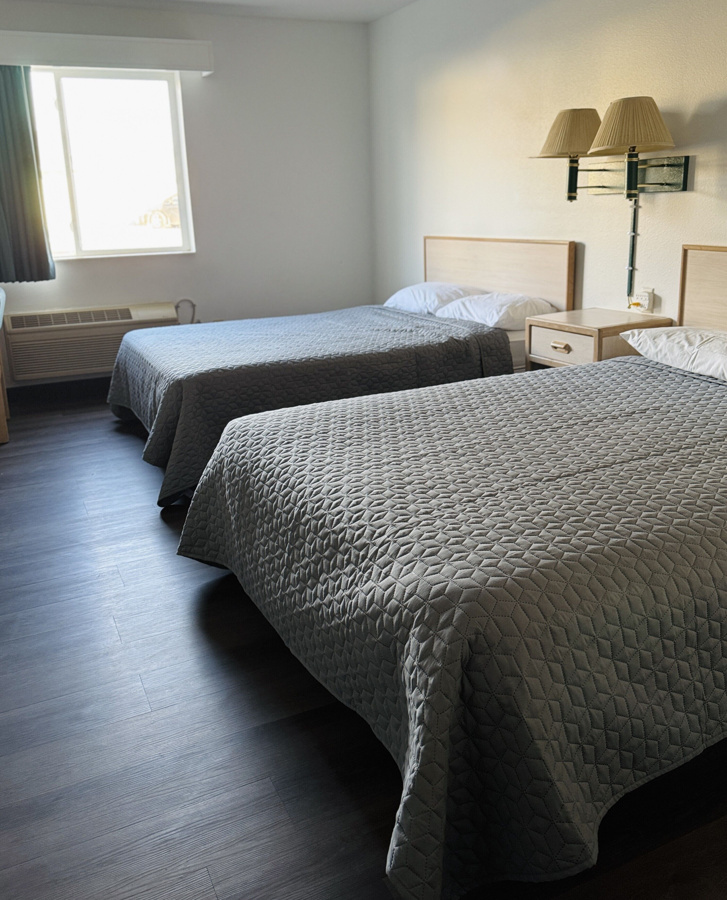 A hotel room with two beds, a nightstand, a lamp, and a window with curtains.