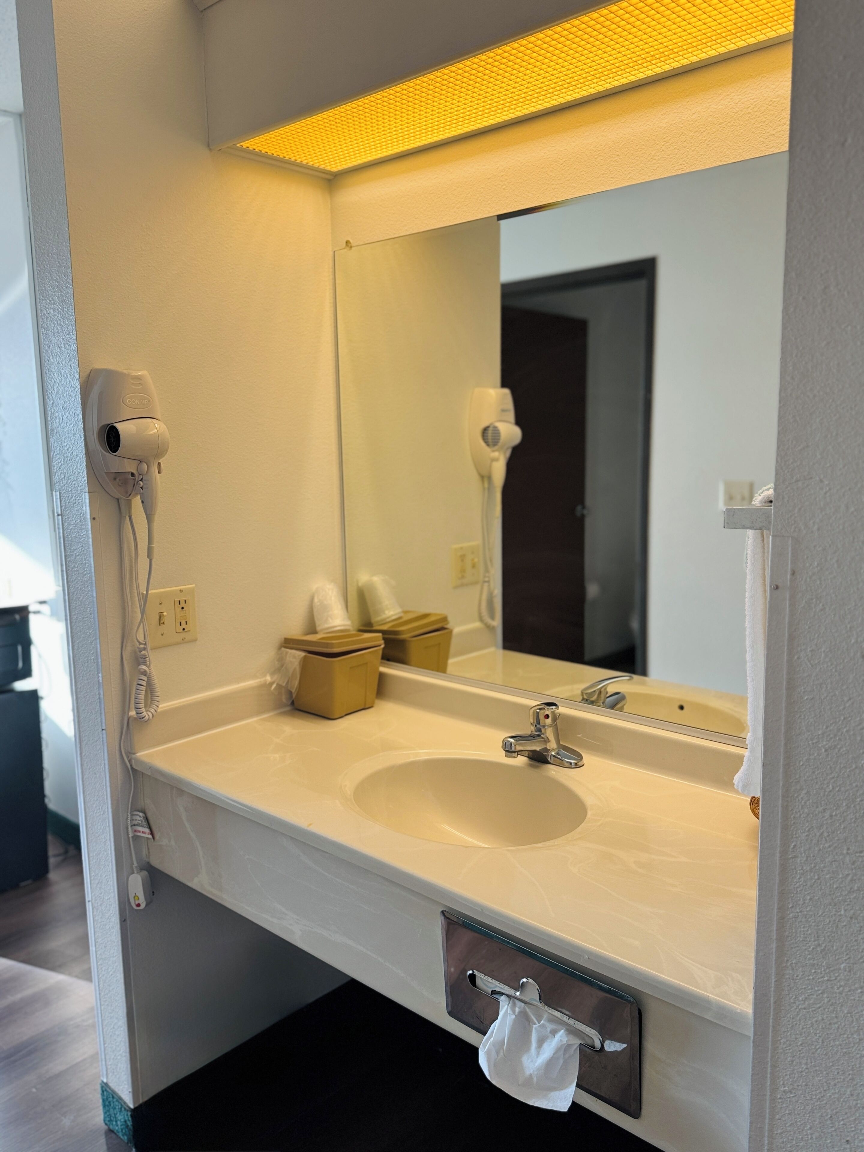 A hotel bathroom with a white sink, a mirror, a hairdryer, and a towel dispenser.