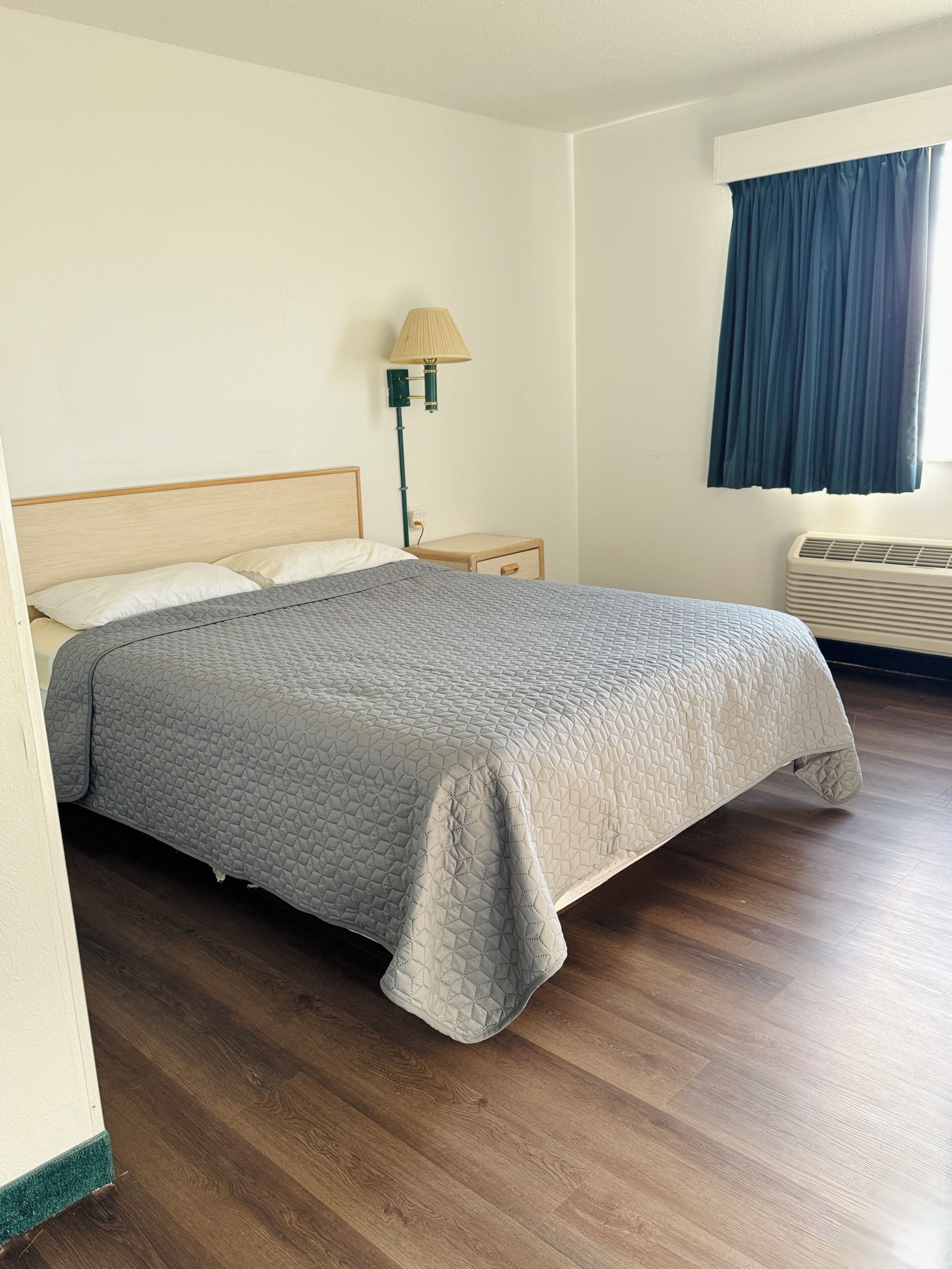 A neatly made bed with a grey quilt, a bedside lamp, a small wooden nightstand, and a window with blue curtains.