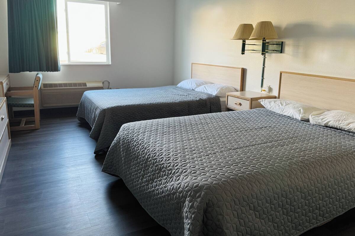 A hotel room with two beds, a desk, a chair, a window with curtains, and a wall-mounted lamp.