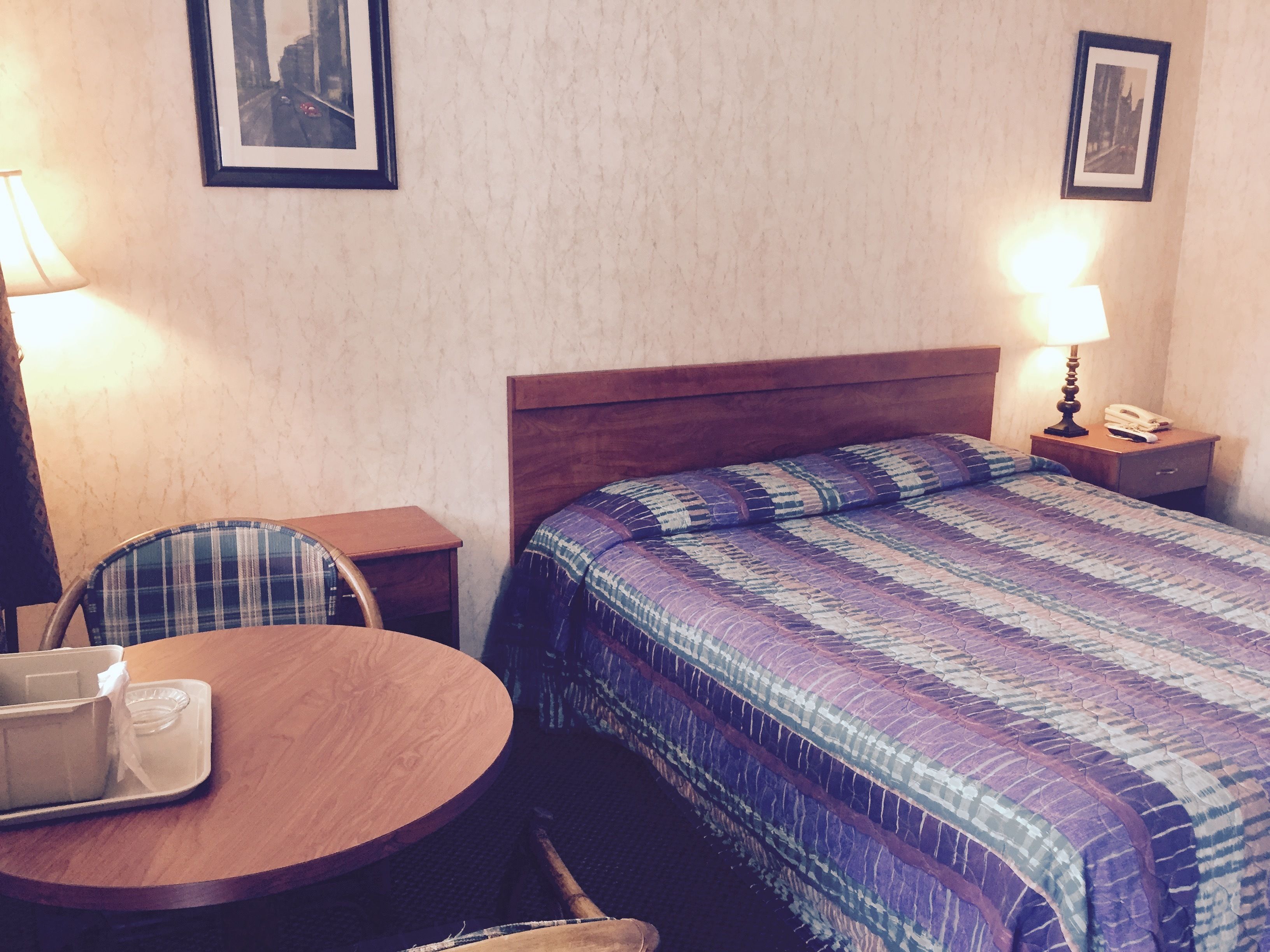 A hotel room with a wooden bed, a round table with a chair, a lamp, and a framed picture on the wall.