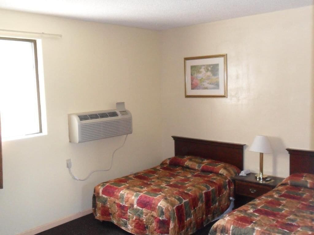 A hotel room with two beds, a wall-mounted air conditioner, a bedside lamp, and a framed picture on the wall.