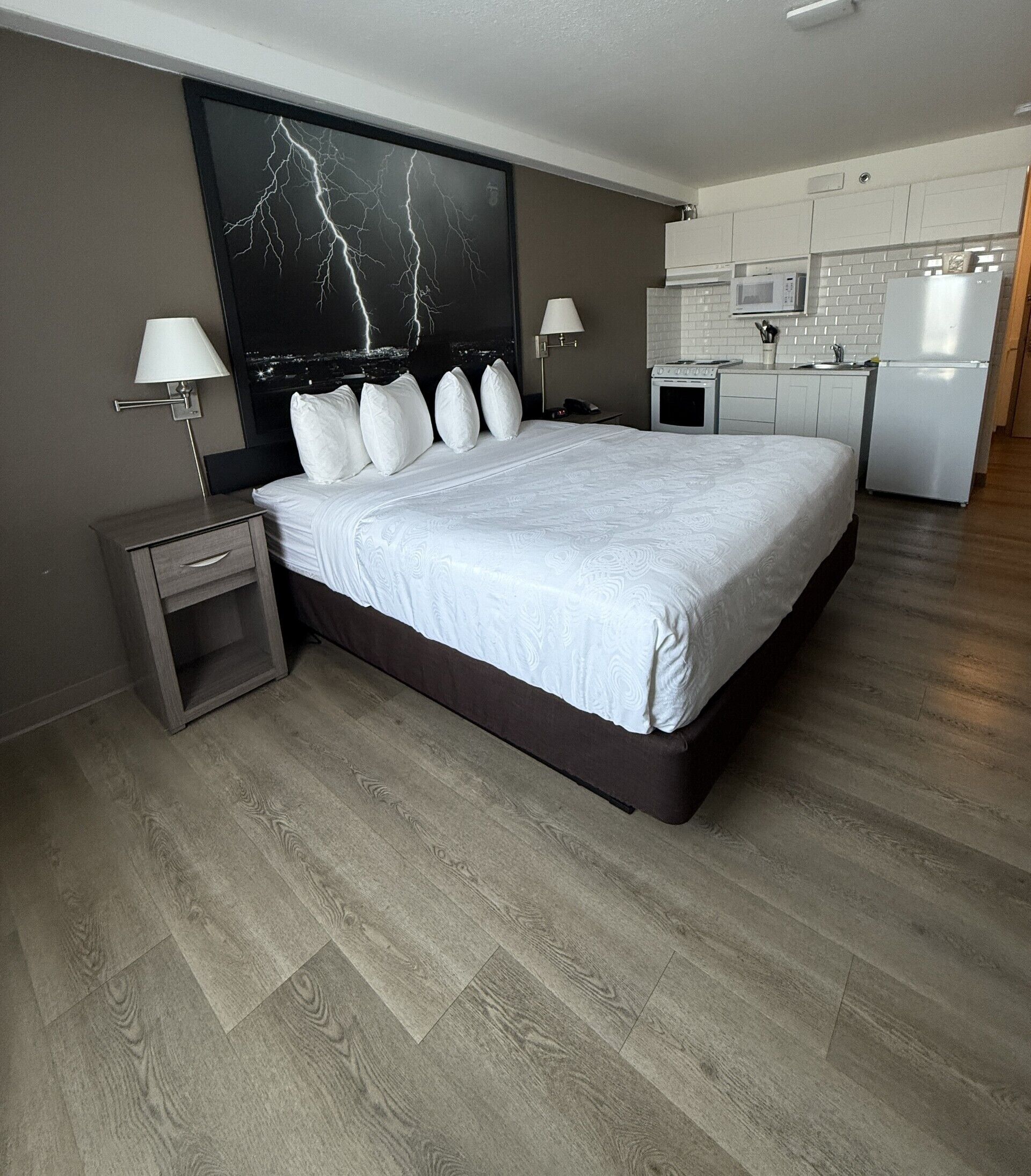 A modern hotel room with a large bed, a nightstand, a desk, a kitchen area, and a view of lightning on the wall.