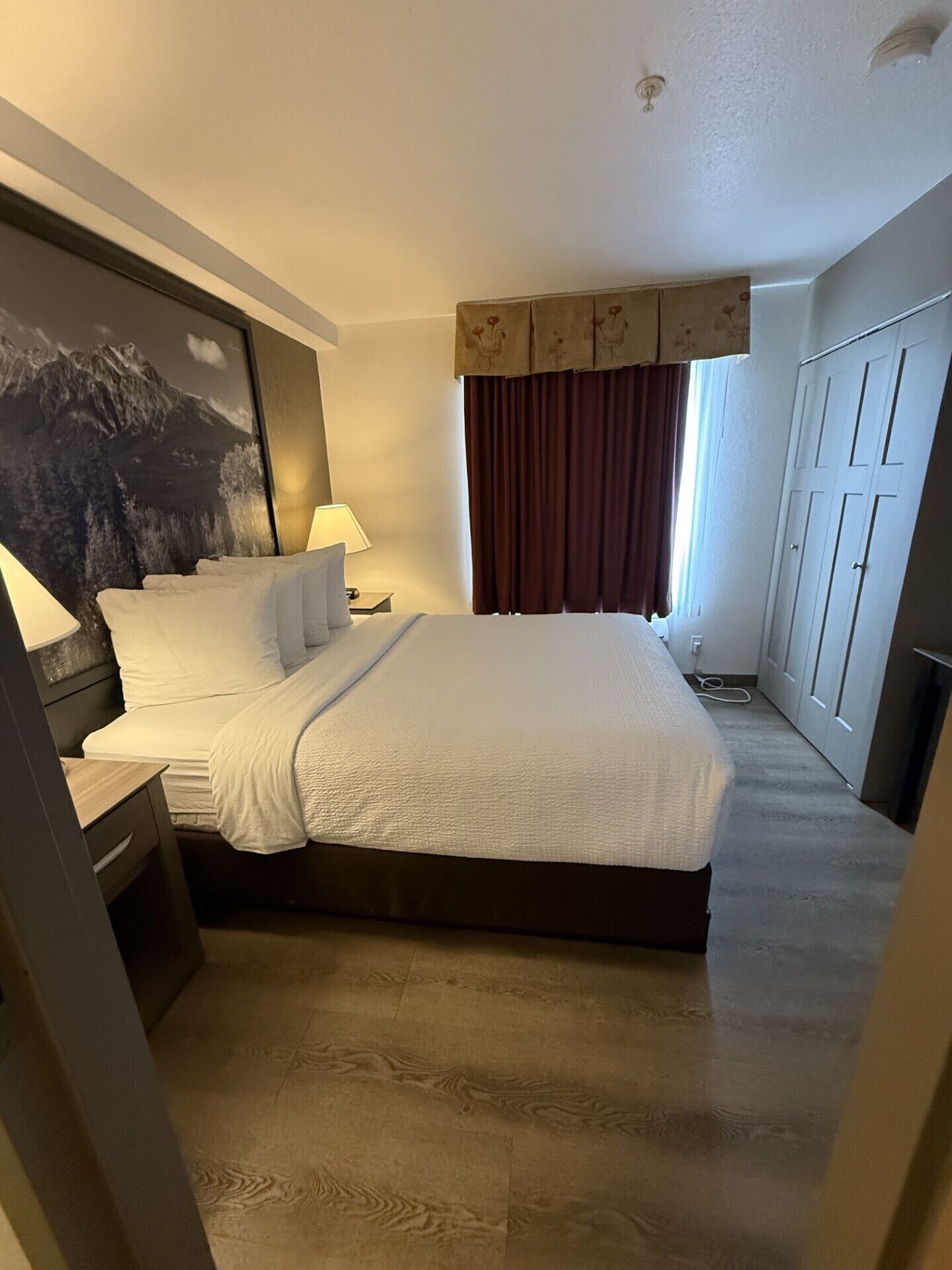 A hotel room with a bed, nightstands, a lamp, a window with curtains, and a wardrobe.