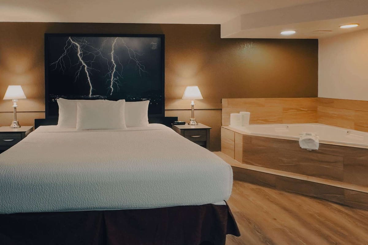 Ein Hotelzimmer mit einem großen Bett, zwei Nachttischlampen, einem am Wand montierten Fernseher, einem Holz-Kopfteil und einer Badewanne.