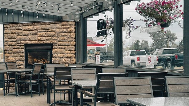 An outdoor seating area with stone walls, a fireplace, and hanging flower baskets. Tables and chairs are arranged under a covered section with a view of the street.