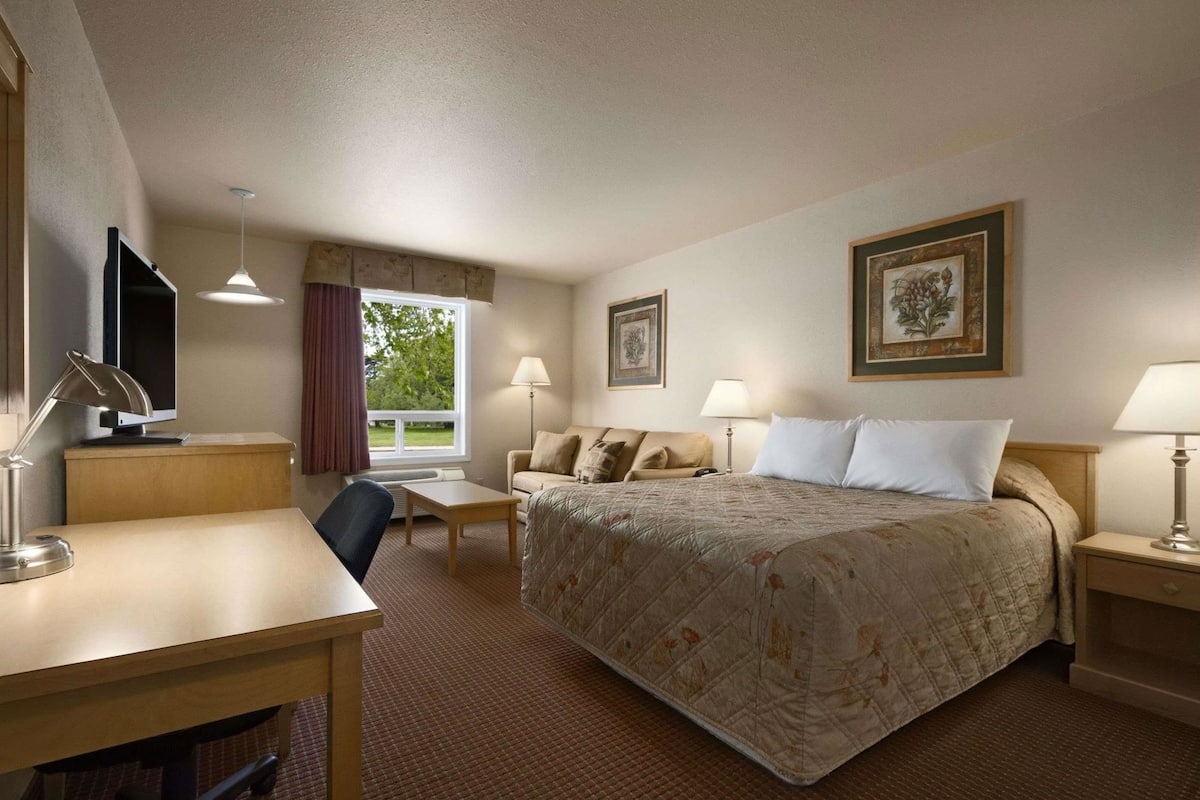 Ein Hotelzimmer mit Bett, Schreibtisch, Stuhl, zwei Lampen, einem Fernseher und gerahmten Bildern an den Wänden.