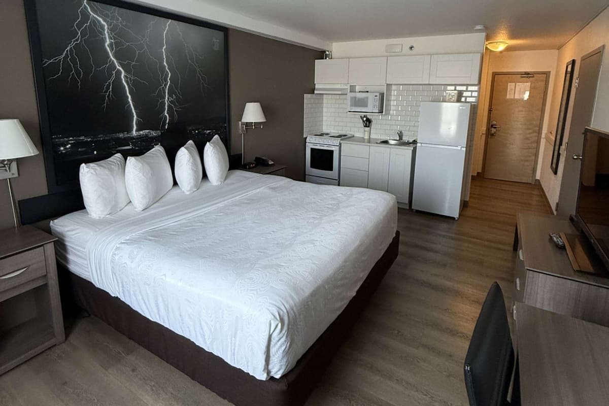 Ein Hotelzimmer mit einem großen Bett, einem Schreibtisch, einem Sessel, einem Fernseher, einer Schreibtischlampe, einer Mikrowelle, einem Kühlschrank und einer Tür zu einem Flur.