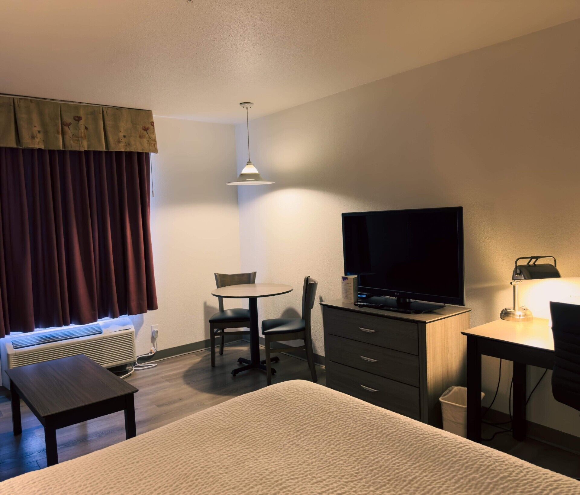A hotel room with a bed, a desk with a computer, a TV, a chair, a small table, and a heater.