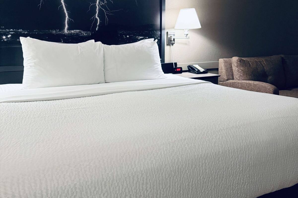 Ein Hotelzimmer mit einem großen Bett, einem Nachttisch mit Lampe und Blick auf eine nächtliche Skyline.