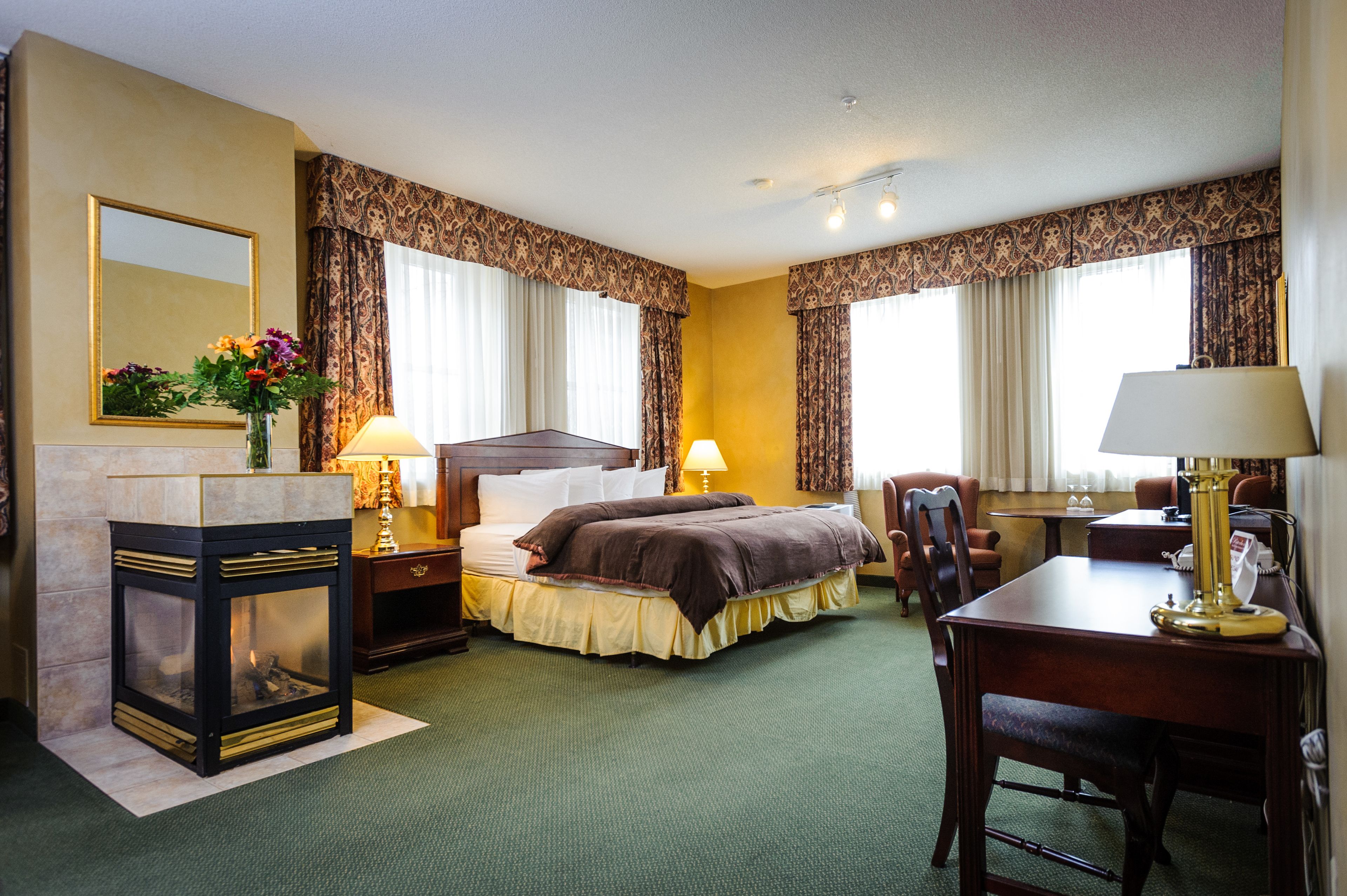 A hotel room with a bed, a desk, a chair, a lamp, a fireplace, and curtains.