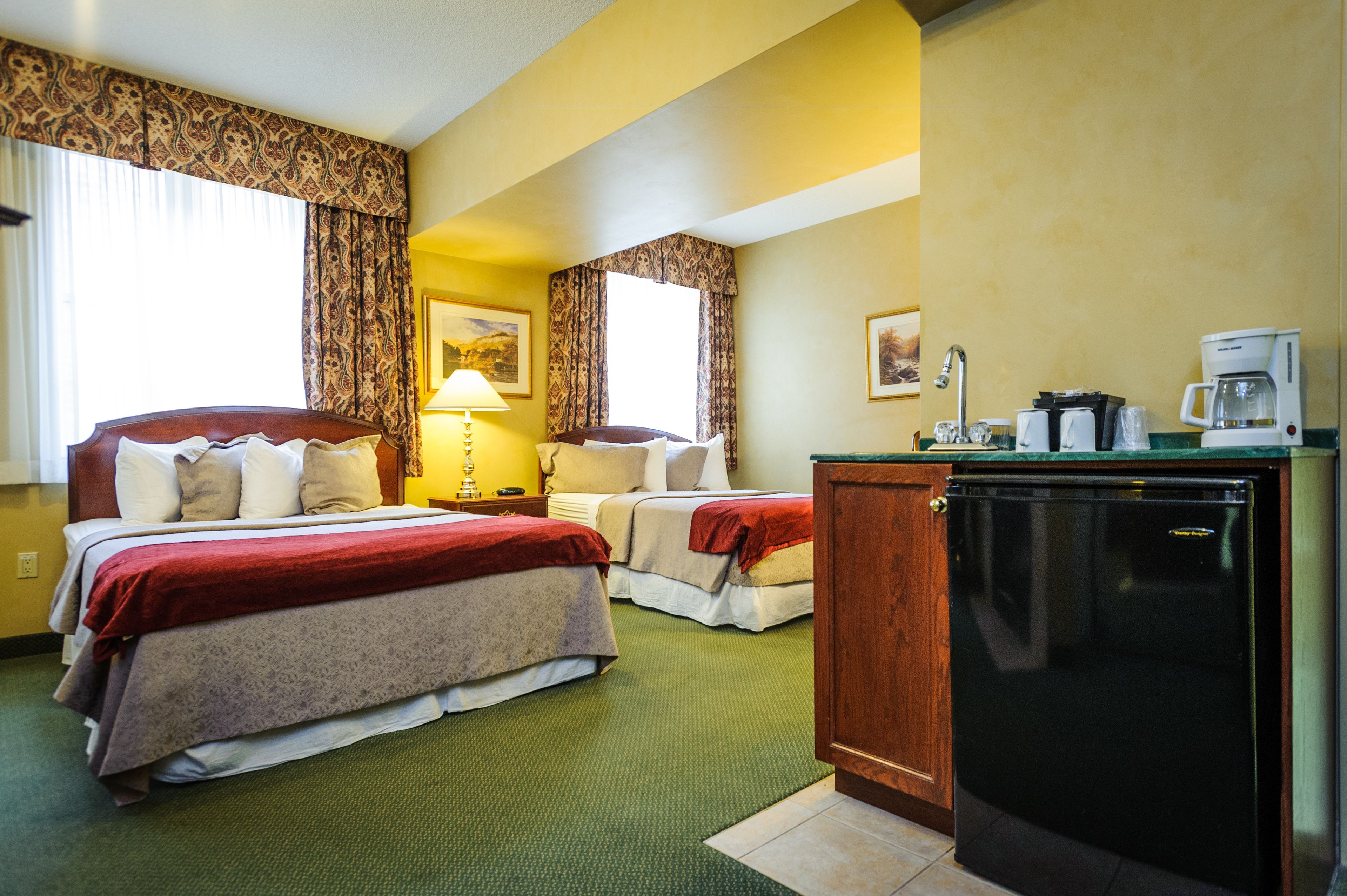A hotel room with two beds, a small kitchenette, and a window with curtains.