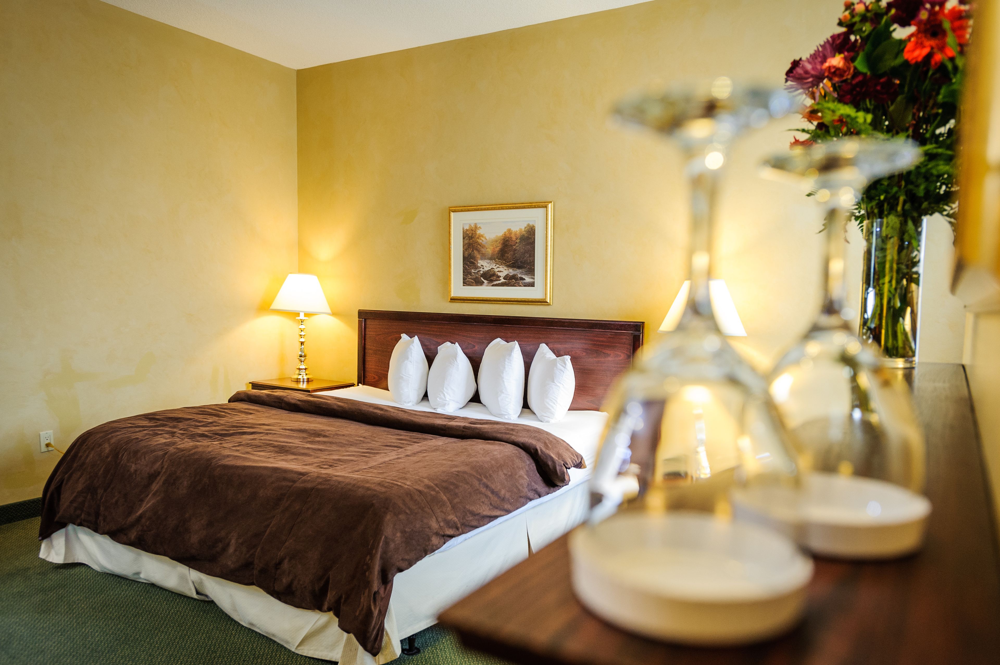 Una habitación de hotel con una cama, una mesita de noche con lámpara, un cuadro en la pared y un jarrón con flores sobre la mesa.