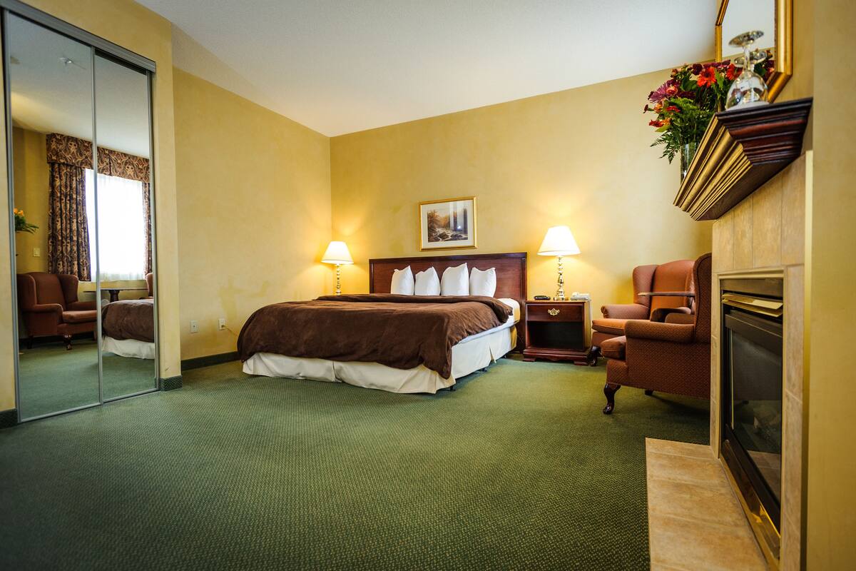 A hotel room with a large bed, two bedside tables with lamps, a chair, a fireplace, and a mirror.
