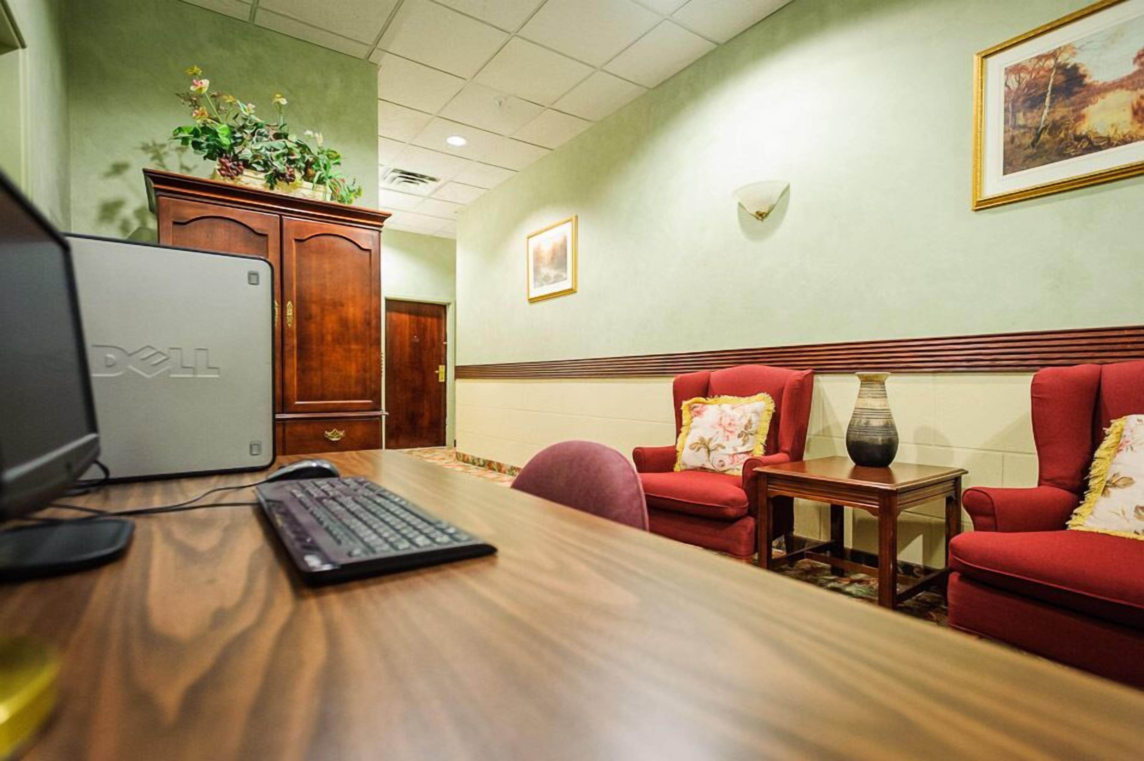 A hotel room with a wooden desk, a Dell computer, a red armchair, and a small table with a vase.