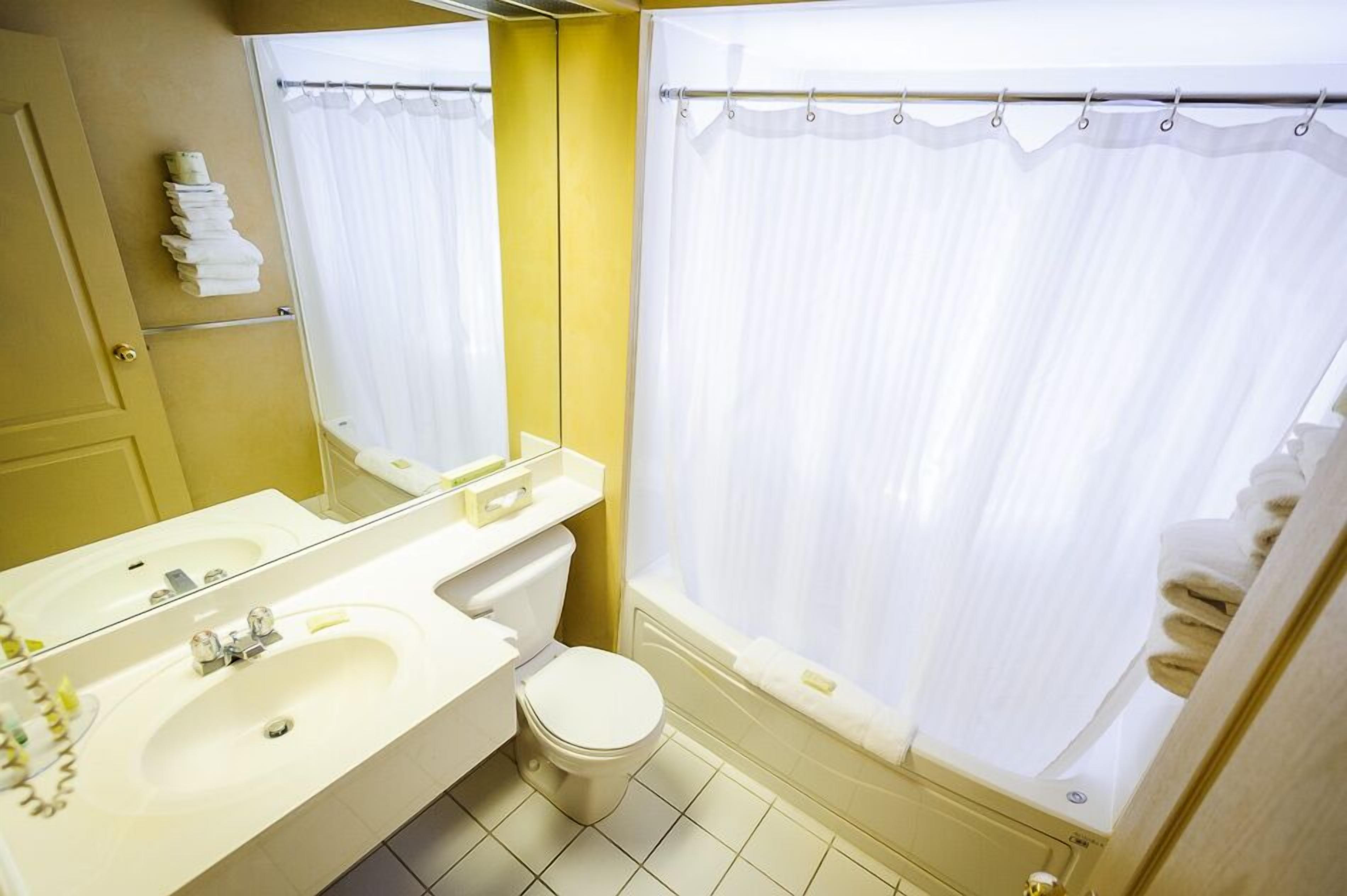 Une salle de bain avec un lavabo, des toilettes et une baignoire blancs, agrémentée d’un mur d’accent jaune et d’un rideau de douche blanc.