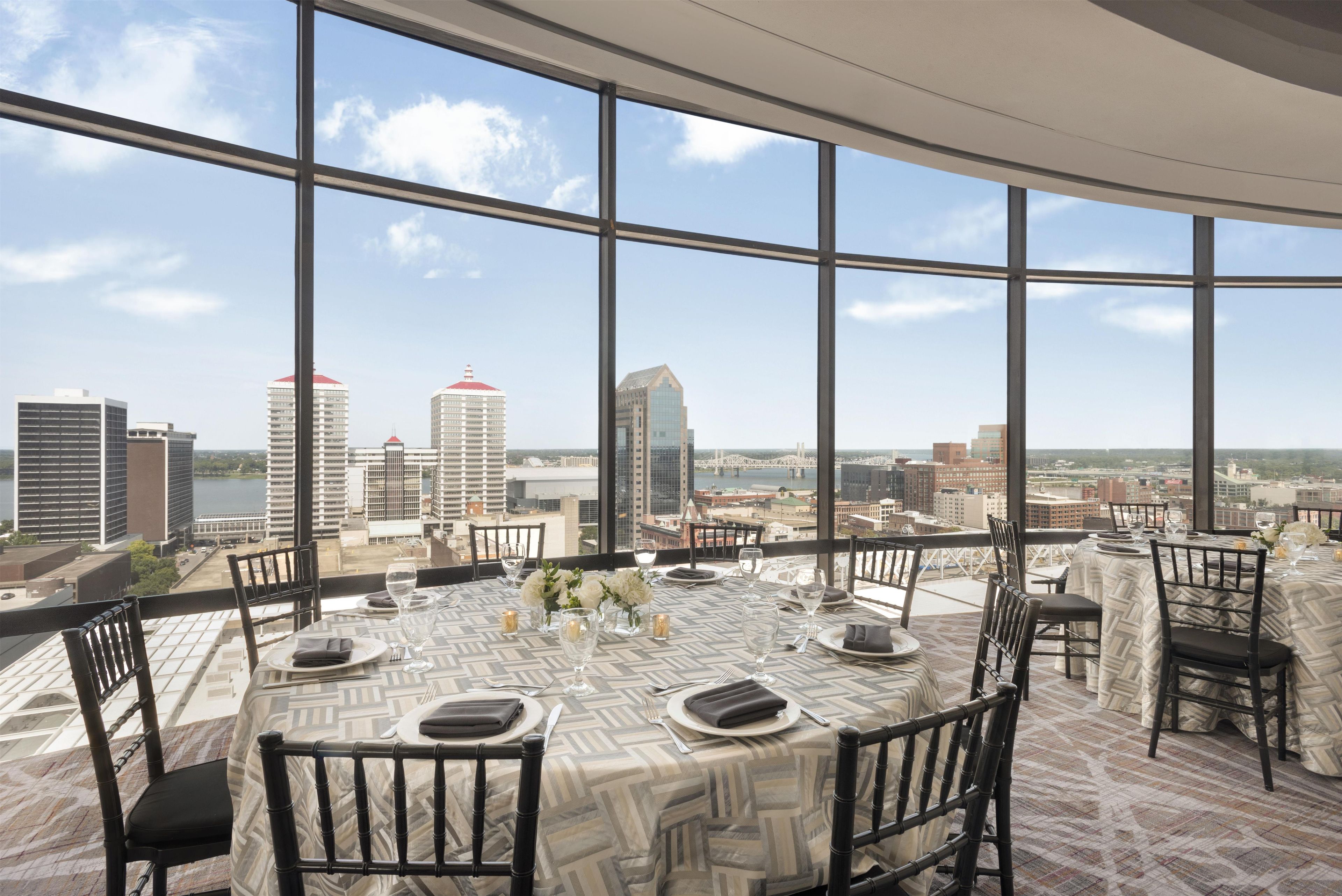 Comedor con vista panorámica de la ciudad, con una mesa redonda dispuesta para cuatro personas, sillas, mantel estampado y un florero con flores.
