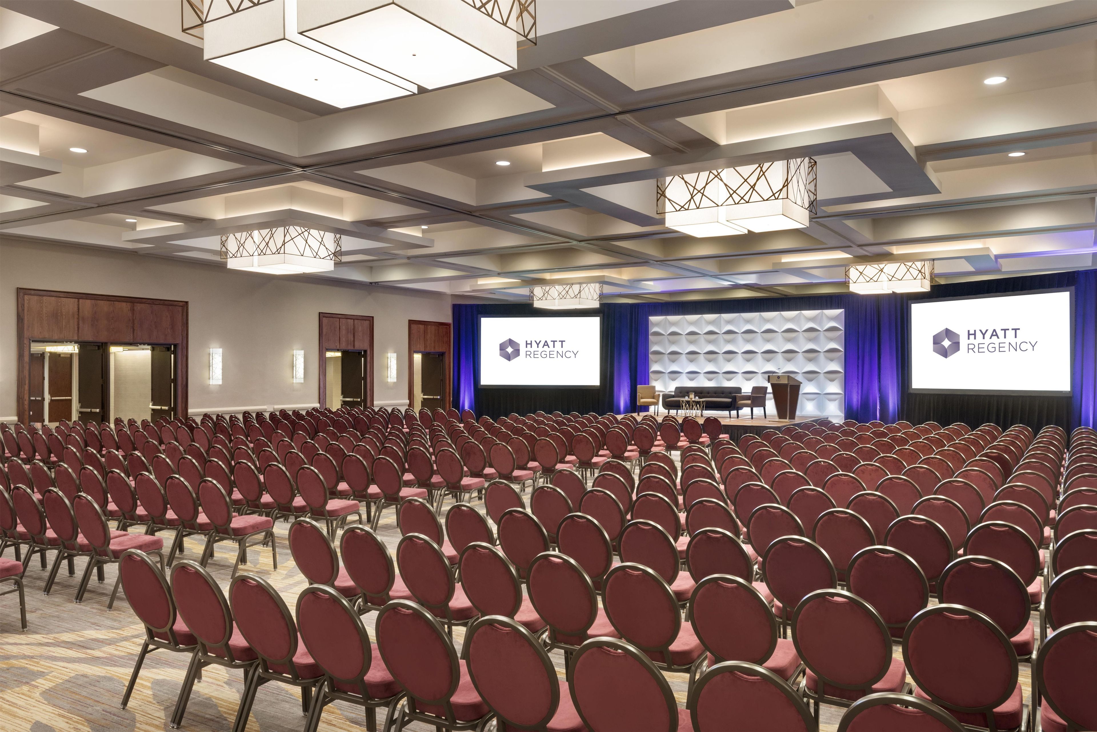 Une salle de conférence avec des rangées de chaises, une scène et un grand écran affichant le logo de l’hôtel Hyatt Regency.
