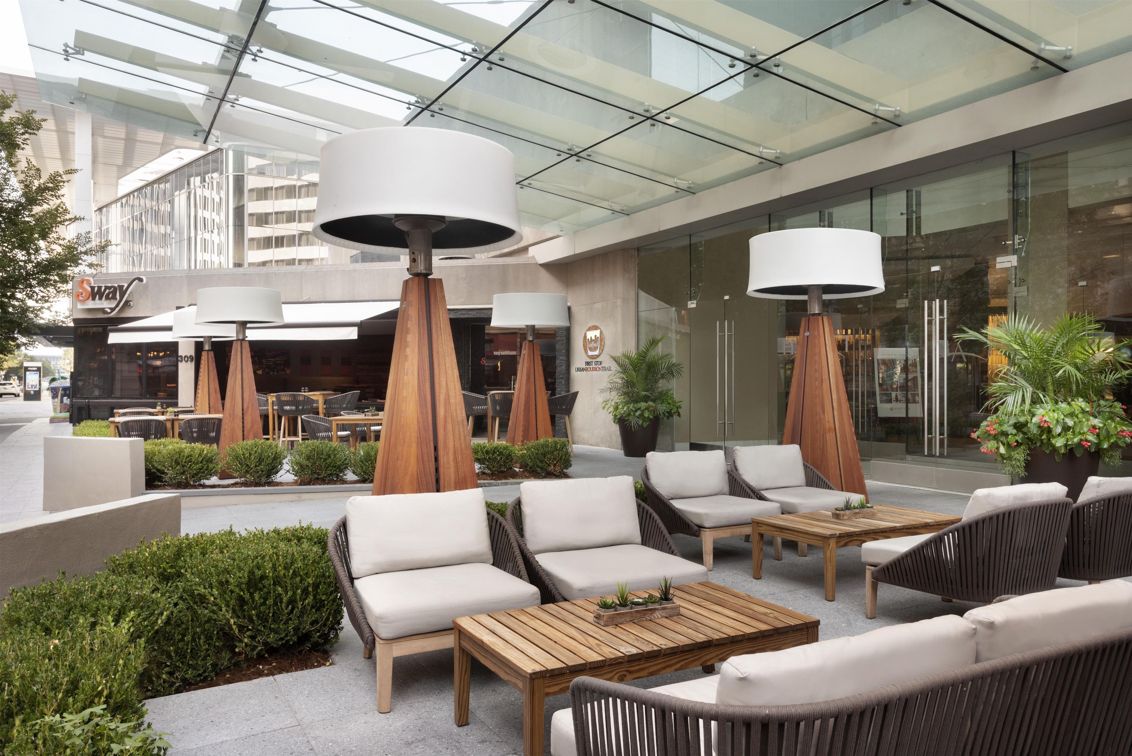 An outdoor seating area with modern furniture and large lamps, a glass ceiling, and a view of the cityscape.
