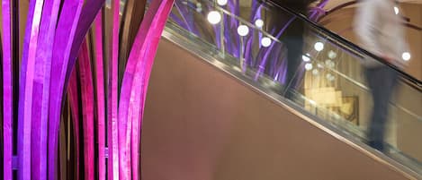 A modern lobby with a curved metallic structure, a seating area with a sofa and a small table, and an escalator in the background.