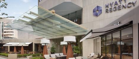 Exterior moderno de un hotel con un área cubierta de vidrio, sillas de lounge al aire libre y un letrero visible que dice 'Hyatt Regency'.