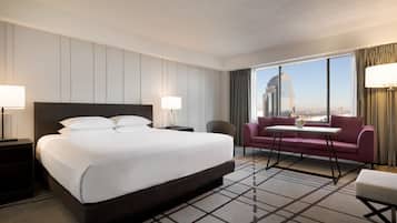 Habitación de hotel con una cama grande, un escritorio, una silla, un sofá y vistas a un paisaje urbano a través de la ventana.