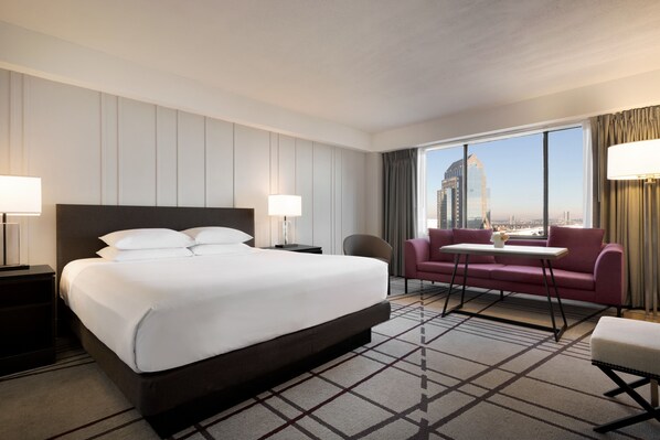 Habitación de hotel con una cama grande, un escritorio, una silla, un sofá y vistas a un paisaje urbano a través de la ventana.
