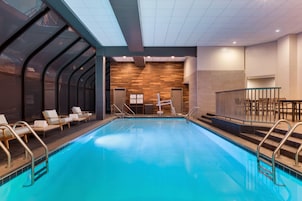 A modern indoor swimming pool with clear glass walls, lounge chairs, and a wooden wall in the background.