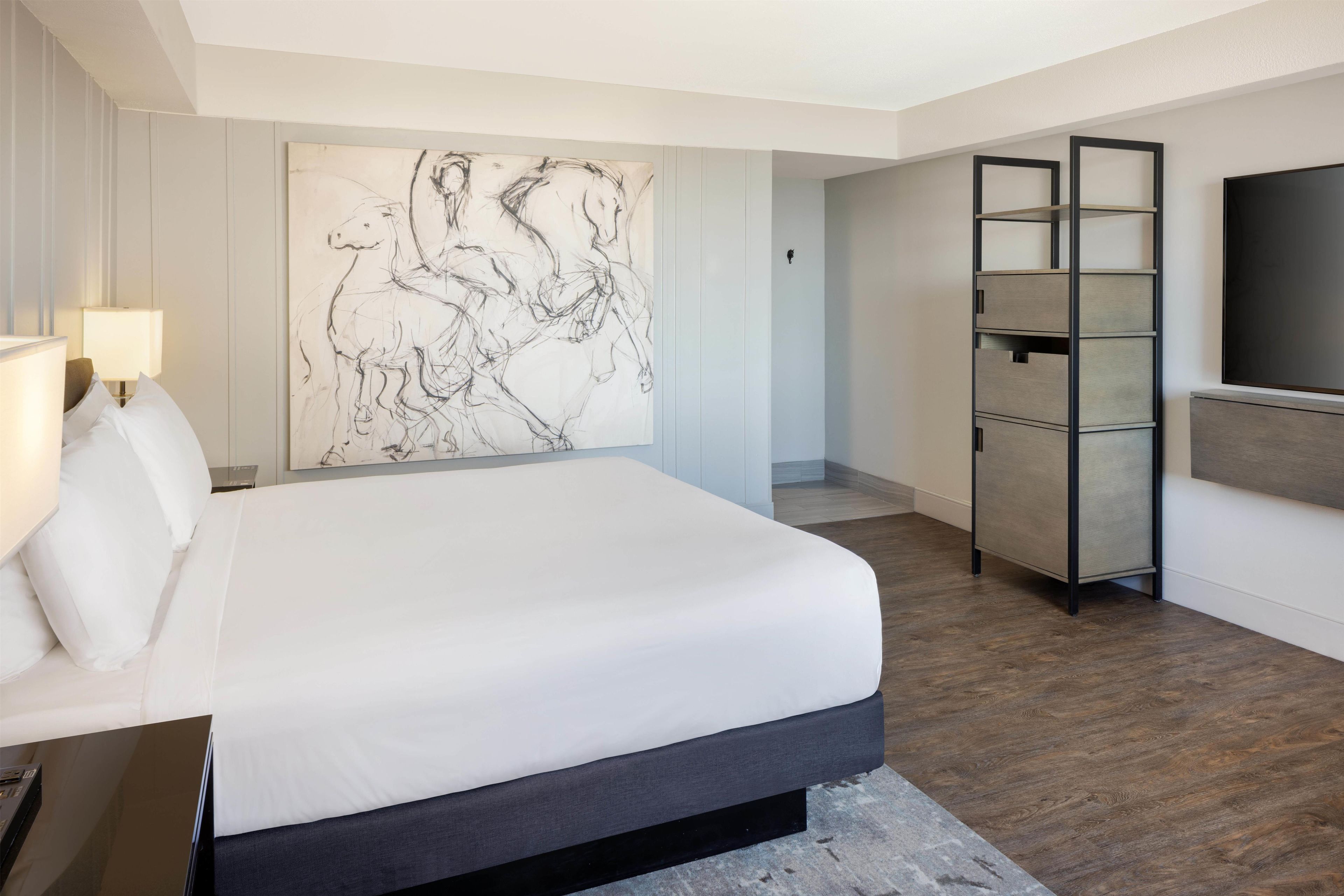 A hotel room with a bed, a wall-mounted TV, a side table with a lamp, and a large abstract painting.