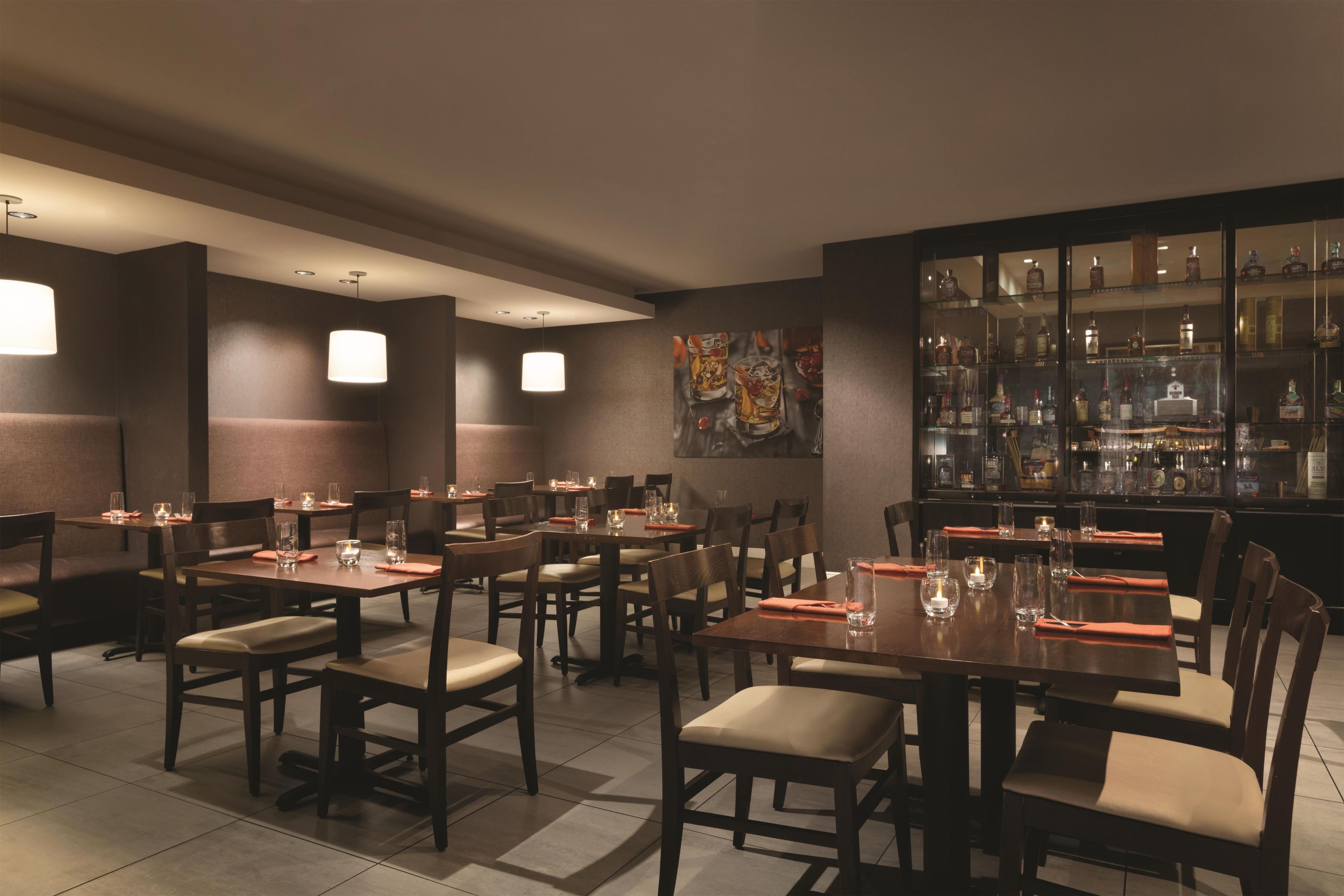 Un restaurante con varias mesas y sillas, una zona de bar con botellas de licor, y un cuadro colgado en la pared.