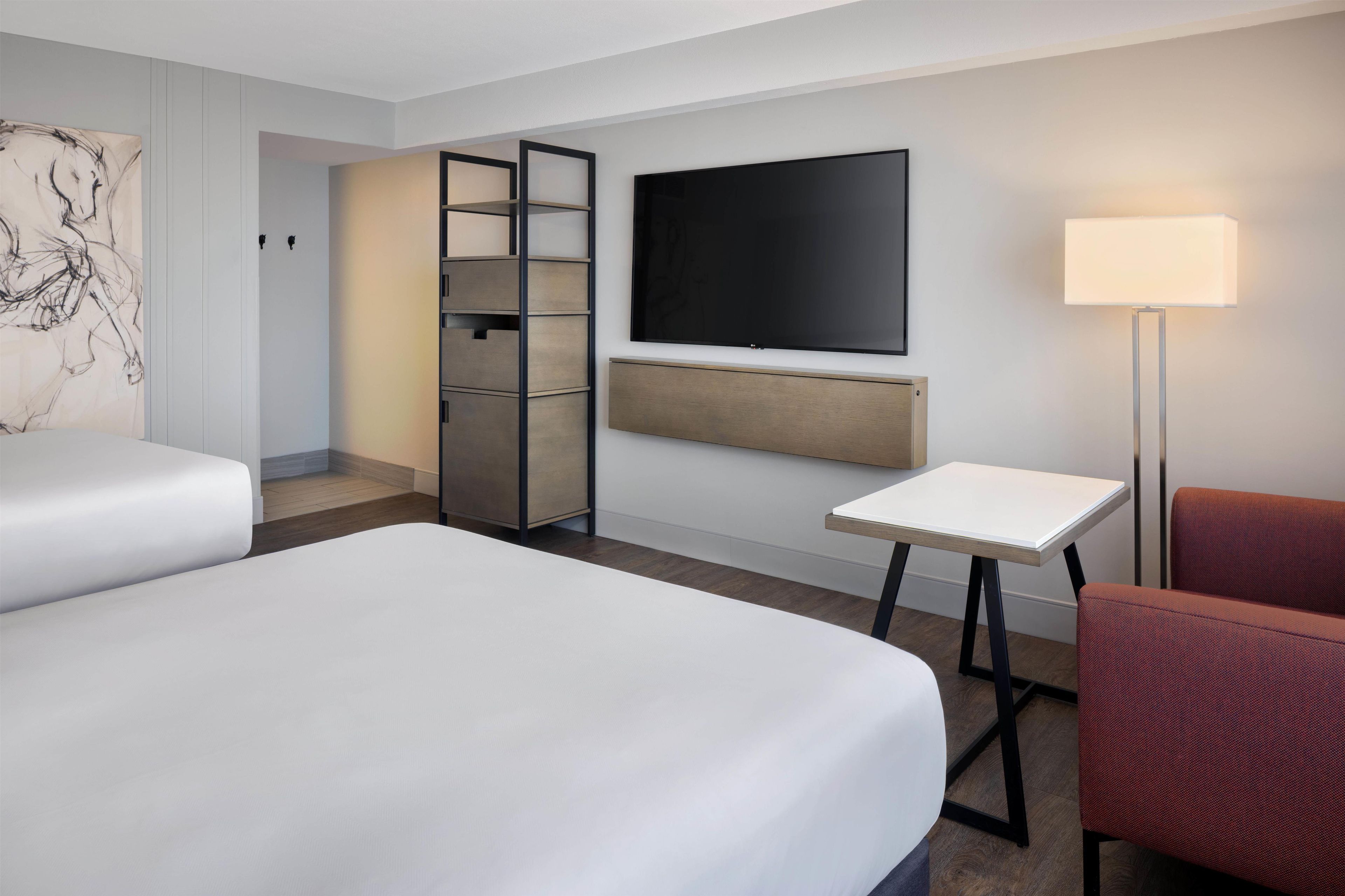 A hotel room with two beds, a desk, a chair, a TV, and a lamp.