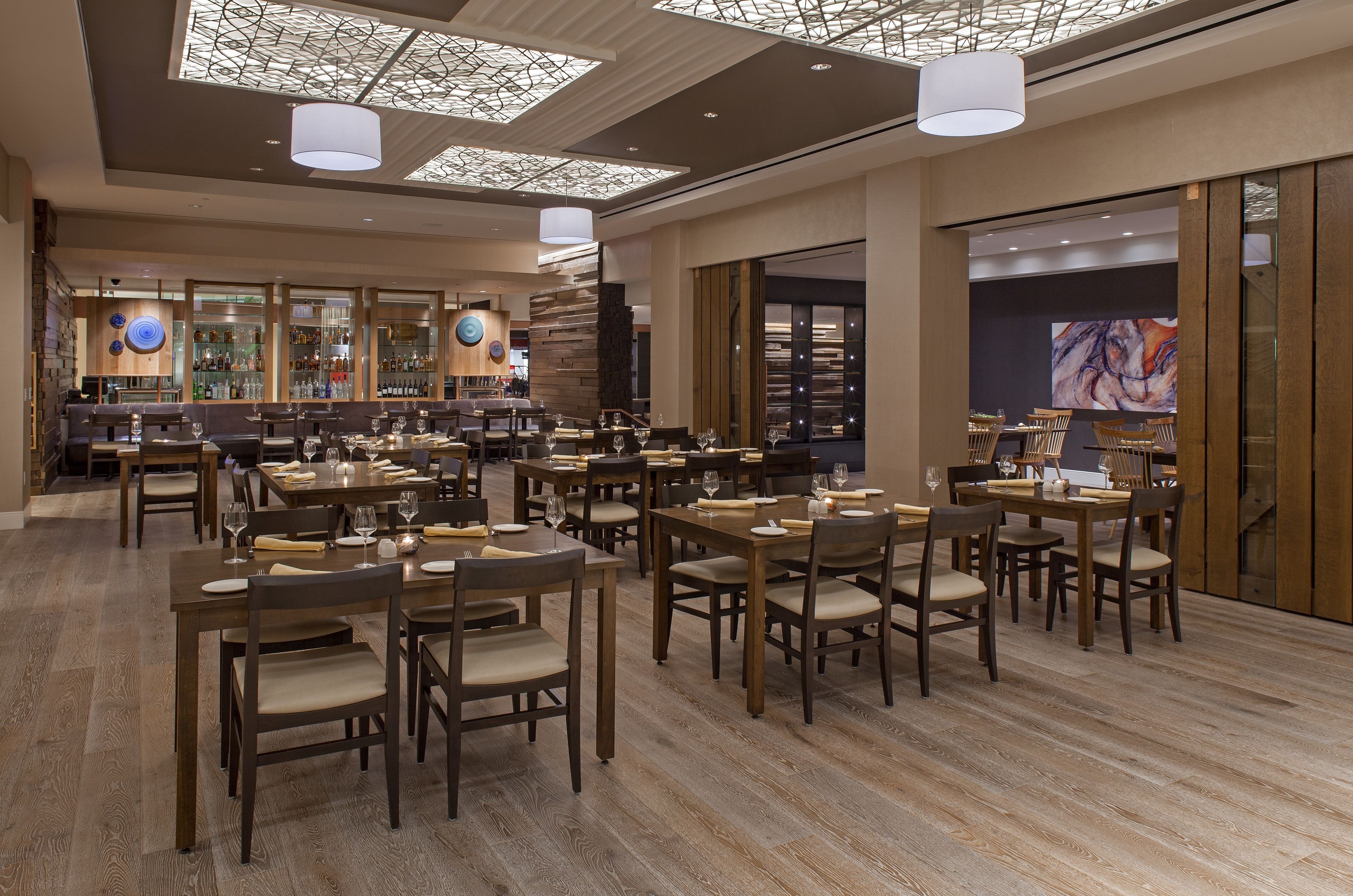 A spacious restaurant with multiple dining tables and chairs, a bar area with shelves of bottles, and a large window with a view.