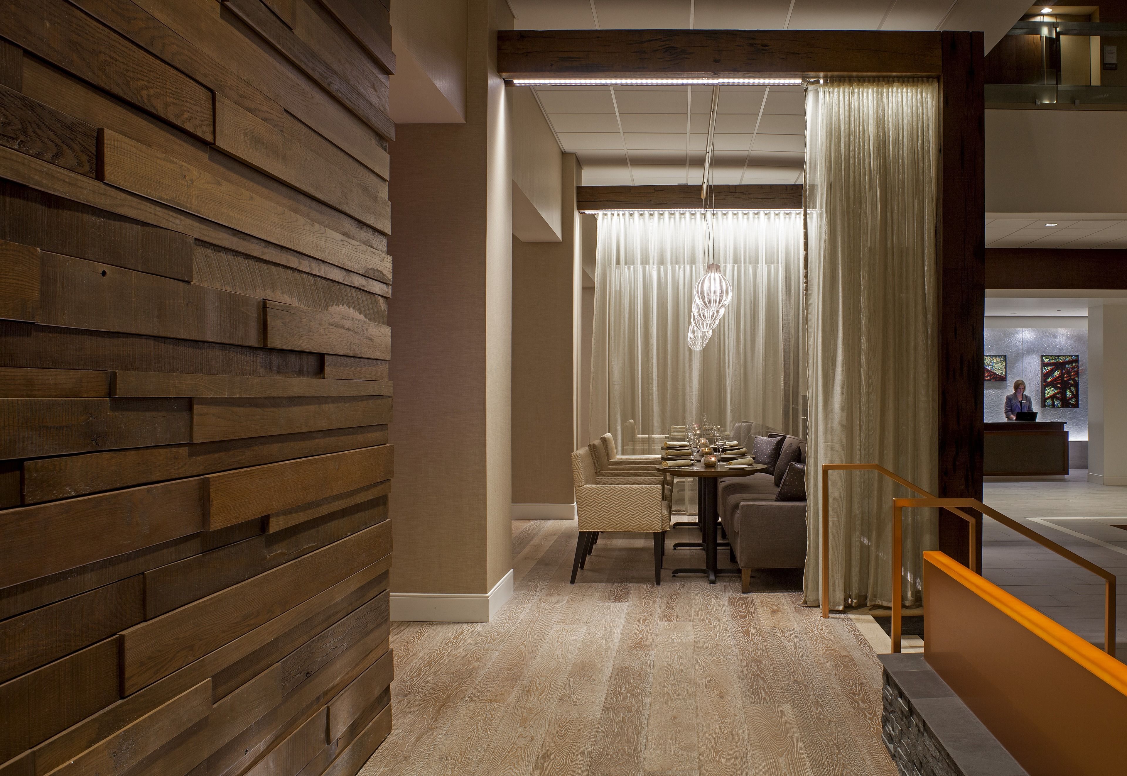 A modern hotel lobby with a wooden wall, a reception desk, and a dining area with a chandelier.