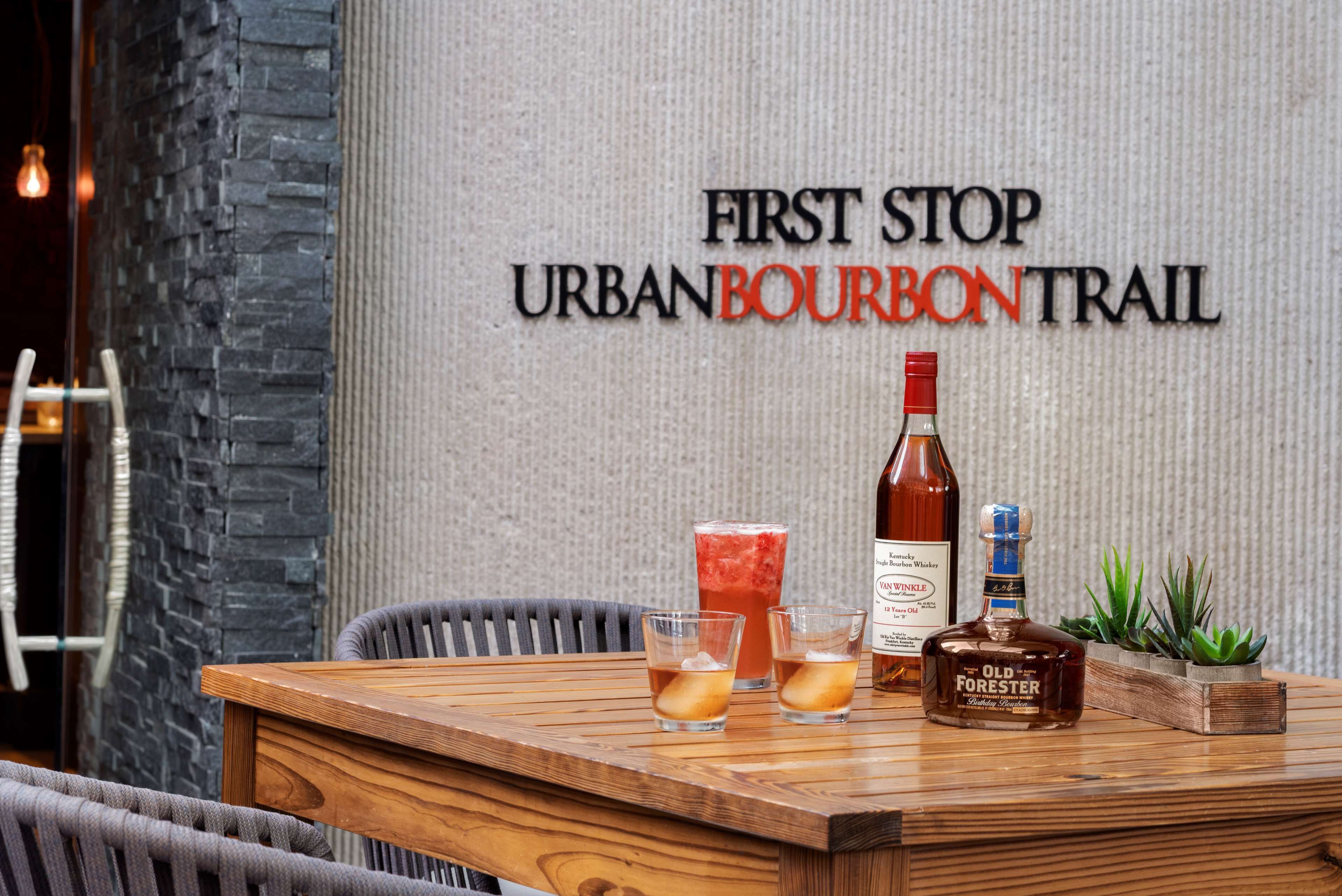 Une table en bois sur laquelle sont posés une bouteille de whisky Old Forester, deux verres remplis de glaçons et un cocktail dans un verre, le tout placé devant un mur sur lequel on peut lire le texte : « FIRST STOP URBANBOURBONTRAIL ».