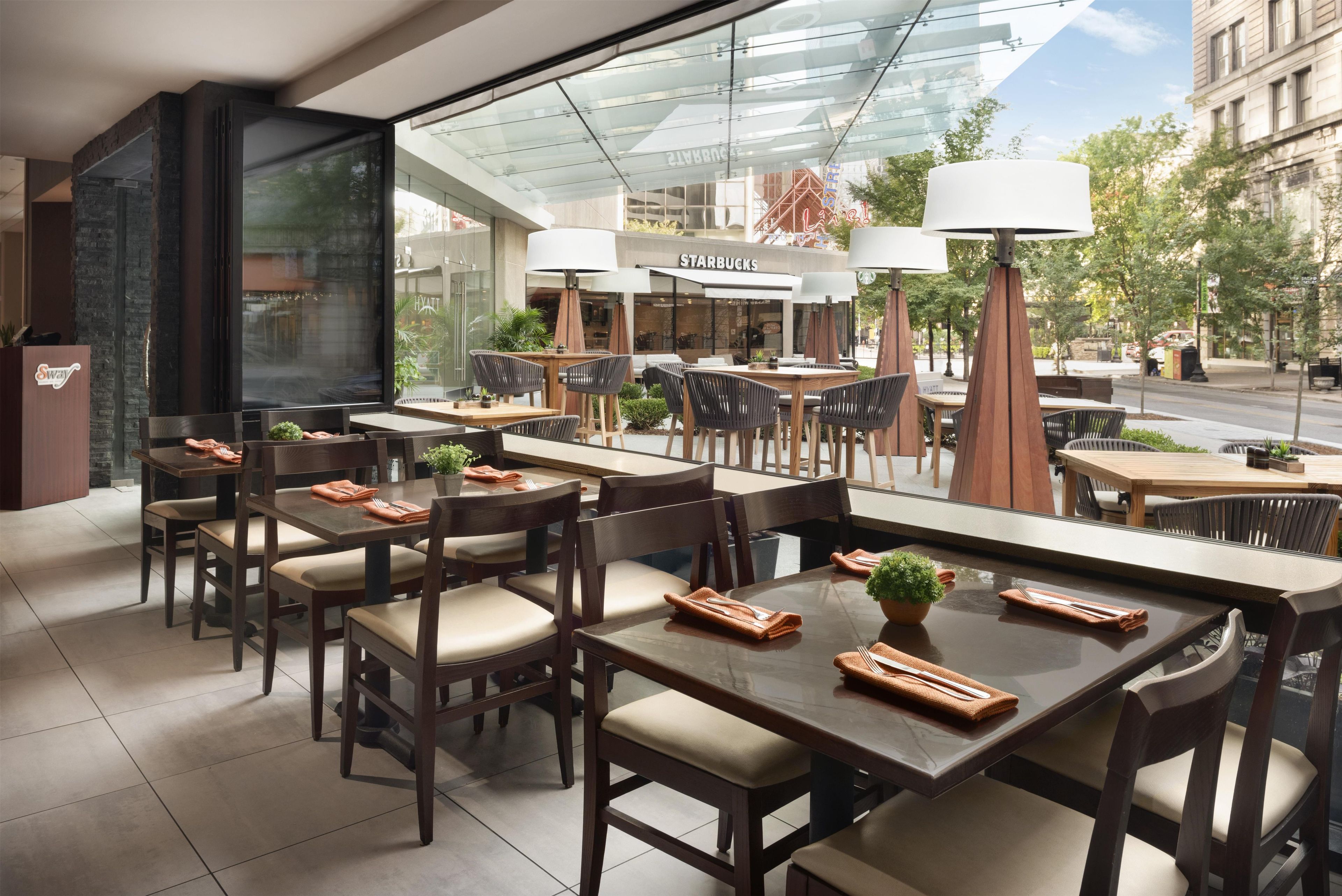 A modern hotel lobby with a Starbucks and Hyatt sign, outdoor seating with tables and chairs, and a glass facade overlooking a street.